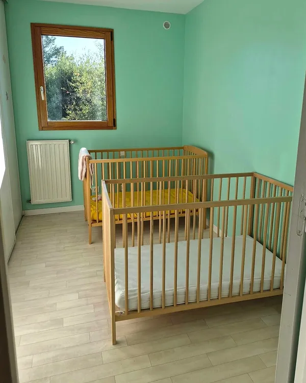 Chambre aux murs vert d’eau avec deux lits à barreaux et une fenêtre sur la verdure